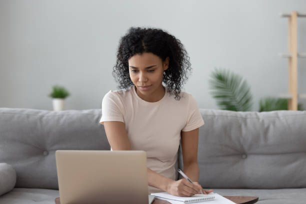 African woman sit on couch at home looks at computer screen holding pen makes notes working doing remote distant job, female studying learning get knowledge using online course self-education concept